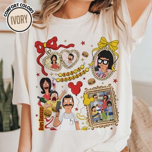 May include: Ivory-colored t-shirt featuring a colorful graphic of characters from Bob's Burgers. The design includes framed portraits, burgers, and the text "Bob's Burgers". The shirt has short sleeves and a crew neck.