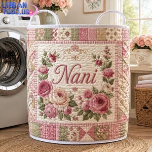 May include: A cylindrical fabric laundry hamper featuring a quilted texture with a patchwork border in shades of pink, cream, and sage green. The front displays the embroidered name Nani surrounded by a floral wreath of pink and cream roses.