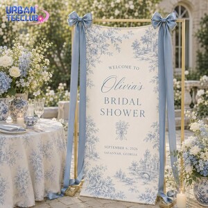 May include: A vertical rectangular bridal shower welcome sign featuring a cream-colored fabric with blue toile floral patterns. The text reads: Welcome to Olivia's Bridal Shower, September 6, 2026, Savannah, Georgia. It is held by a gold frame with blue ribbons.