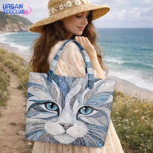 May include: A tote bag featuring a detailed, artistic illustration of a cat face with striking blue eyes. The cat's fur is rendered in shades of blue, gray, and white with intricate, swirling leaf-like patterns throughout the design.