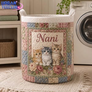 May include: A cylindrical quilted fabric laundry basket featuring a patchwork pattern in pastel shades of pink, blue, and green. The front displays a central rectangular panel with the name Nani and three kittens surrounded by floral accents.