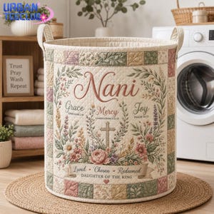 May include: A cylindrical quilted fabric laundry hamper featuring a patchwork border in muted green, pink, and cream tones. The center displays the name Nani, the words Grace, Mercy, Joy, and a cross, surrounded by floral illustrations and text.