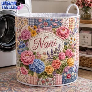 May include: A cylindrical fabric laundry hamper featuring a quilted patchwork design with various floral patterns in pink, blue, and yellow. The center displays a white oval patch with the embroidered name Nani surrounded by colorful flowers.
