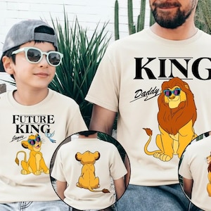 May include: Beige t-shirts with lion graphics. One shirt says "KING Daddy" with a lion wearing sunglasses. The other says "FUTURE KING" with a lion and a bird. The back of the shirts also have lion graphics.