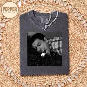 May include: Dark gray Comfort Colors t-shirt featuring a black and white image of a person biting a golf ball on a green lawn. A silver chain necklace is draped across the shirt. The shirt is folded on a woven, tan rug.