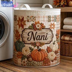 May include: A cylindrical fabric laundry hamper featuring a quilted patchwork design in autumn colors. The exterior displays pumpkins, fall leaves, and flowers, with a central rectangular plaque bearing the name Nani in dark brown script.