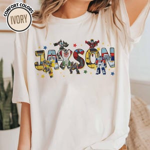 May include: Ivory-colored t-shirt featuring the name "Jaxson" in large, colorful letters with Transformers characters. The design includes stars and the words "Comfort Colors" and "Ivory" in a circle.