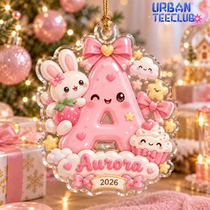 May include: A clear acrylic Christmas ornament shaped like the letter A in pink, featuring a white bunny holding a strawberry, a cupcake, bows, stars, and clouds. The name Aurora and the year 2026 are printed on a banner at the bottom of the ornament.