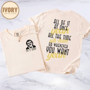 May include: Two ivory-colored t-shirts. One shirt features a black and white image of a man with the text "DO IT LADY!". The other shirt has the text "ALL OF IT AT ONCE YEAH ALL THE TIME UH-HUH OR WHENEVER YOU WANT YEAH" in black and yellow.