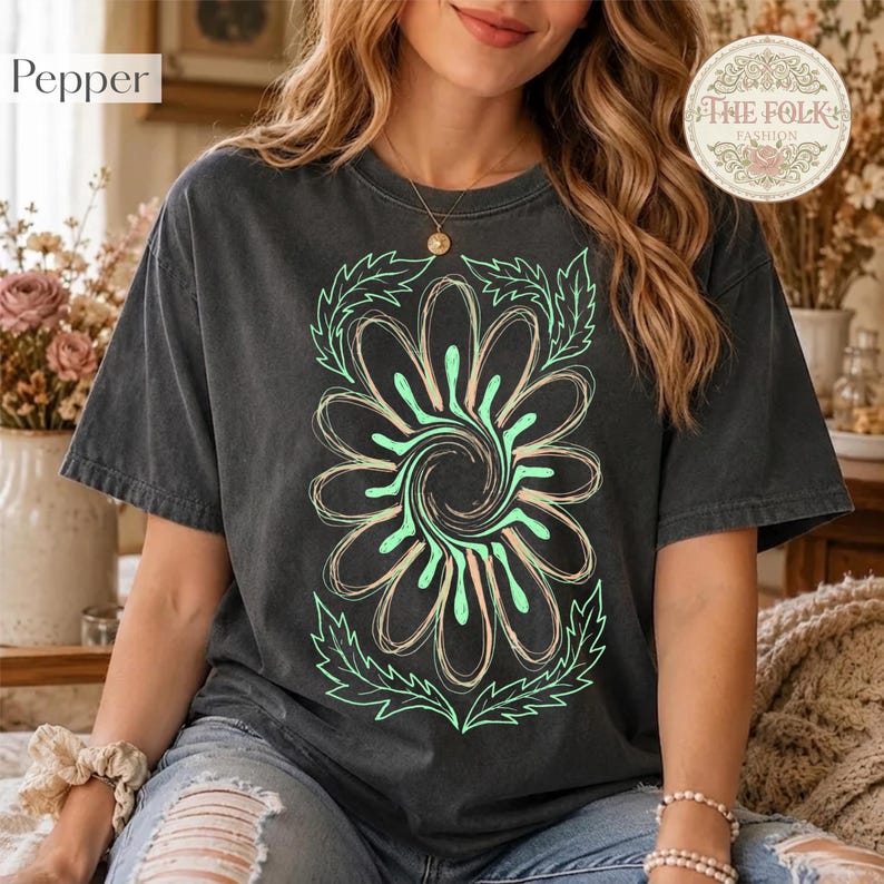 May include: A charcoal gray short-sleeve t-shirt featuring a large, central graphic of a stylized flower with swirling petals in shades of light green and peach, outlined in thin lines, with leafy accents at the top and bottom of the design.