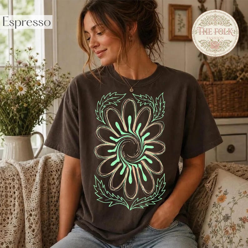 May include: A dark brown short-sleeve t-shirt featuring a large, central graphic of a stylized flower with swirling petals in light green and cream outlines. The shirt has a crew neckline and a relaxed fit, showcasing a bold floral design.