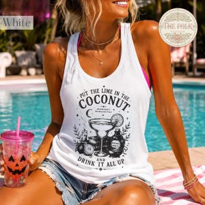 Puede incluir: Una camiseta de tirantes blanca con espalda nadadora que presenta un estampado gráfico negro de una copa de margarita, una rodaja de lima, una botella etiquetada como Witches Spirits y el texto Put the lime in the coconut and drink it all up.