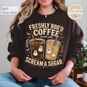 Könnte beinhalten: Ein dunkelbraunes Sweatshirt mit Rundhalsausschnitt und einem Grafikdruck von zwei Eiskaffeebechern mit Geisterillustrationen, Spinnenweben und Fledermäusen. Der Text lautet FRESHLY BOO'D COFFEE served with SCREAM & SUGAR in cremefarbener Schrift.