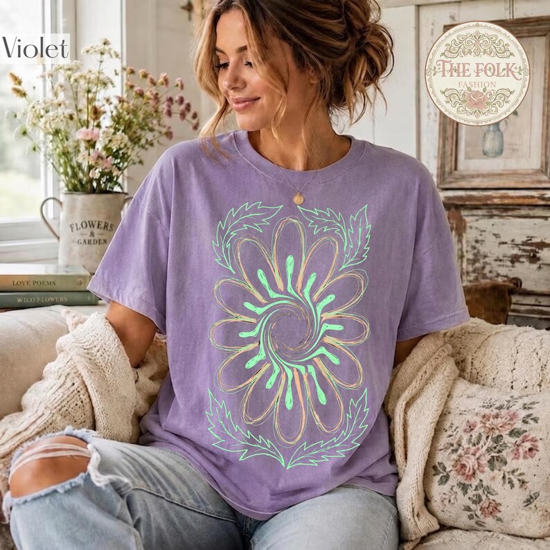 May include: A light purple short-sleeve t-shirt featuring a large, central graphic of a stylized flower with swirling petals in shades of light green and yellow, outlined in thin lines, with leafy accents at the top and bottom of the design.
