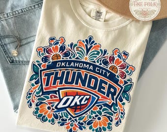 May include: Ivory-colored Oklahoma City Thunder t-shirt with a colorful floral design. The shirt features the team name and logo in blue, orange, and white. The shirt is made of a soft material and is a comfortable fit.