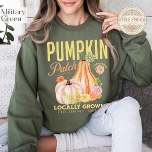 Könnte beinhalten: Ein militärgrüner Rundhals-Pullover mit einem Grafikdruck, der den Text PUMPKIN PATCH LOCALLY GROWN CIDER CORN MAZE PUMPKIN zeigt. Das Design enthält Illustrationen von zwei Kürbissen, verschiedenen Herbstblumen und Blattwerk in Orange-, Rosa- und Gelbtönen.