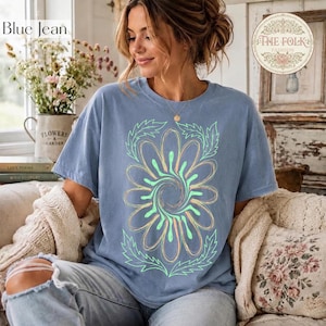 May include: A blue short-sleeved t-shirt featuring a large, central graphic of a stylized flower with swirling petals in light green and peach tones, surrounded by leafy outlines. The shirt has a crew neckline and a relaxed, casual fit.