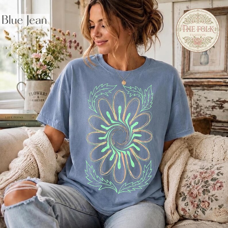 May include: A blue short-sleeved t-shirt featuring a large, central graphic of a stylized flower with swirling petals in light green and peach tones, surrounded by leafy outlines. The shirt has a crew neckline and a relaxed, casual fit.