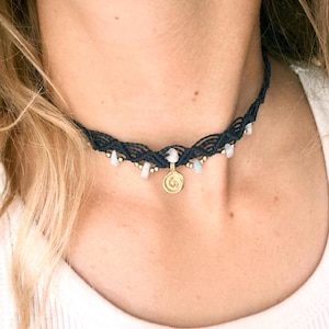 Puede incluir: Un collar gargantilla de macramé azul marino con pequeñas cuentas blancas y doradas. Un colgante dorado con un diseño en espiral cuelga del centro. El collar se lleva alrededor del cuello.