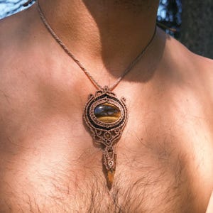 May include: A macrame necklace with a large oval tiger's eye stone pendant. The pendant is intricately woven with brown cord and accented with dark blue and black details. A smaller, elongated crystal hangs below the stone, suspended from a woven cord.