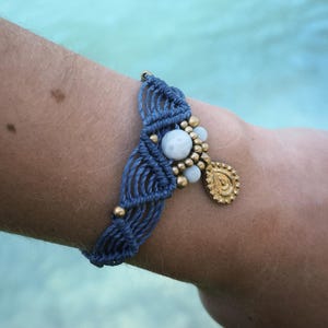 Könnte beinhalten: Ein handgefertigtes blaues Makramee-Armband mit goldenen Perlen und einem goldenen Tropfenanhänger. Das Armband hat eine zentrale Ansammlung weißer und goldener Perlen. Der Hintergrund ist eine verschwommene türkisfarbene Farbe.