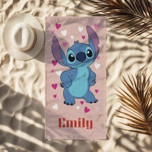 Puede incluir: Una toalla de playa rosa con un personaje de dibujos animados azul, orejas grandes y corazones rosas y blancos. El nombre "Emily" est&aacute; impreso en rojo. Se ve un sombrero blanco para el sol. Sobre arena.