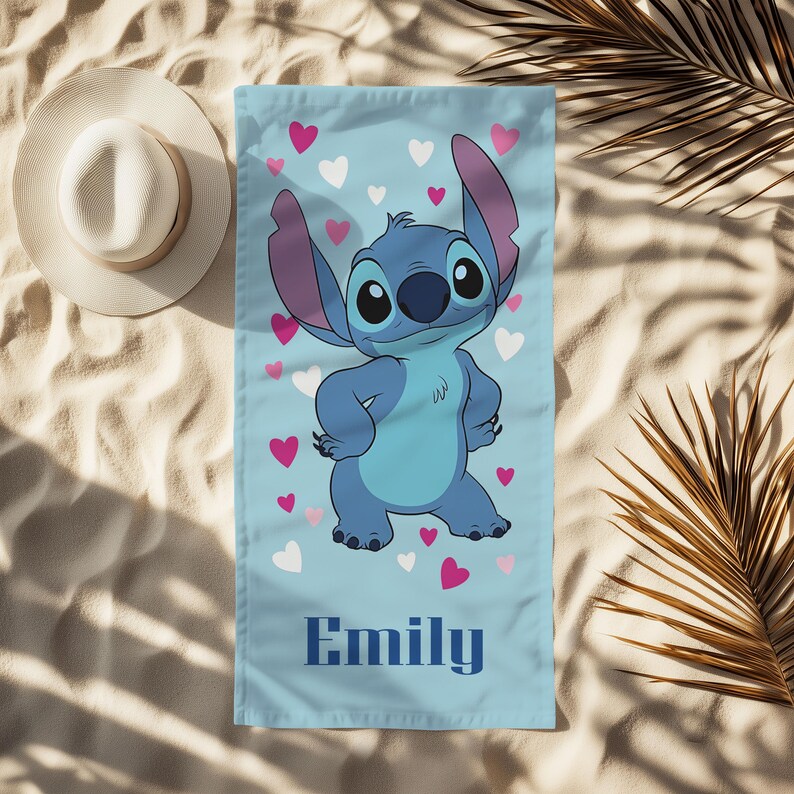 Puede incluir: Toalla de playa azul claro con un personaje de dibujos animados con orejas grandes y una expresi&oacute;n amigable, rodeado de corazones rosas y blancos. El nombre "Emily" est&aacute; impreso en azul marino en la parte inferior. Un sombrero blanco para el sol es visible en la esquina superior izquierda.
