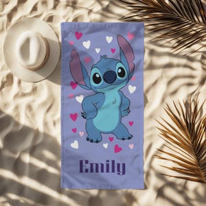 Puede incluir: Una toalla de playa lavanda con una imagen de dibujos animados de Stitch, rodeada de corazones rosas y blancos. La toalla tiene el nombre "Emily" impreso en una fuente morada oscura. Se ve un sombrero para el sol blanco.