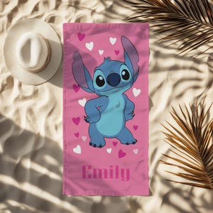 Puede incluir: Toalla de playa rosa con el personaje Stitch de Lilo & Stitch, rodeado de corazones rosas y blancos. La toalla tiene el nombre "Emily" impreso en rosa. Un sombrero de sol blanco es visible en el fondo.