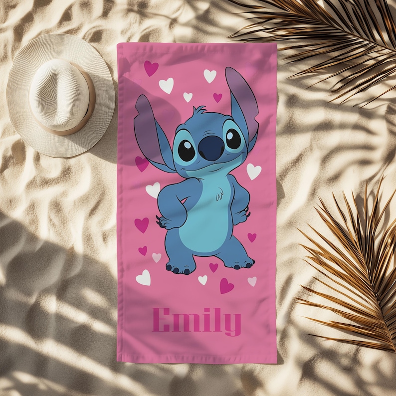 Puede incluir: Toalla de playa rosa con el personaje Stitch de Lilo & Stitch, rodeado de corazones rosas y blancos. La toalla tiene el nombre "Emily" impreso en rosa. Un sombrero de sol blanco es visible en el fondo.