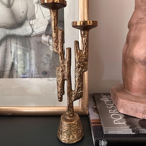 May include: A bronze-colored, two-candle candelabra with an abstract, textured design. The candelabra has a wide base and two holders for candles. The design is reminiscent of a cactus. The candelabra is displayed on a dark surface.