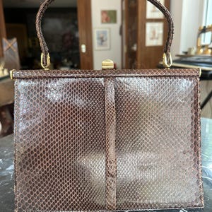 May include: A structured brown handbag with a textured snakeskin pattern, featuring a single top handle, a gold-toned metal frame, and a central gold-toned clasp closure. The bag has a rectangular shape with a vertical strip running down the centre.