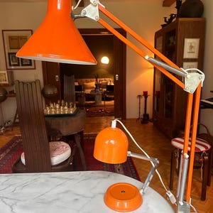 May include: An orange desk lamp with a cone-shaped shade and adjustable arm. The lamp has a smaller, matching orange light. The base is silver. The lamp is on a white marble table.
