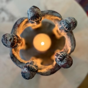 May include: A top-down view of a circular, textured candle holder made of dark, weathered material with a distressed finish. Five rounded, protruding elements form the rim, surrounding a single, glowing, warm-toned tea light candle.