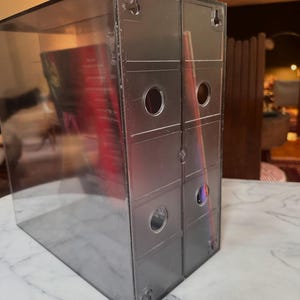 May include: A clear, rectangular display case with a gray, metallic back. The case has a transparent front and sides, with circular holes and hanging hardware on the back. The case is designed to hold items, with a red object visible inside.