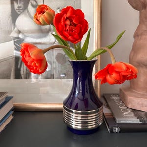 May include: A vibrant floral arrangement featuring bright red tulips in a navy blue vase with silver accents. The vase sits on a dark surface, with a framed black and white artwork and a bust sculpture in the background.