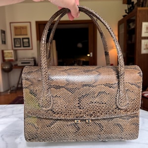 May include: A white ceramic mug with a black handle and the text "But First, Coffee" in a bold, black font. The mug is set against a blurred background, suggesting a cozy setting.