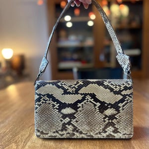 Może przedstawiać: Prostokątna torebka ze wzorem skóry węża w odcieniach beżu, brązu i czerni. Torba ma pasujący pasek i jest trzymana przez rękę. Torba leży na drewnianym stole.