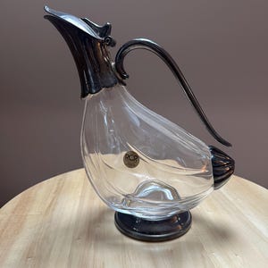 May include: Clear glass jug with a silver-coloured handle and spout. The jug has a unique shape and sits on a dark base. The jug has the letters "RCR" on a sticker. A decorative piece for serving beverages.