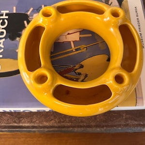 Peut inclure: Un cendrier en céramique jaune vif de forme circulaire. Le cendrier comporte quatre ouvertures circulaires et une ouverture centrale. Il est posé sur une affiche vintage.