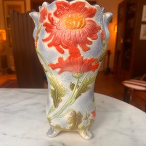 Może przedstawiać: Ceramiczny wazon z jasnoniebieskim szkliwionym tłem oraz wypukłymi, trójwymiarowymi reliefami przedstawiającymi duże, żywe pomarańczowe kwiaty chryzantemy i zielone liście. Wazon ma falisty brzeg i stoi na trzech małych nóżkach.