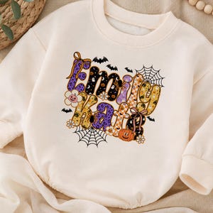 May include: A cream-coloured crewneck sweatshirt featuring a central graphic with the name Emily Kate in large, stylised letters. The design incorporates Halloween motifs including orange pumpkins, black spiders, spiderwebs, bats, and flowers.