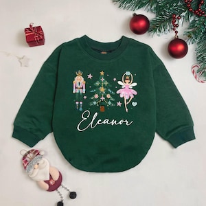 May include: A dark green sweatshirt featuring a Christmas-themed design. The design includes a nutcracker, a Christmas tree, and a ballerina. The name "Eleanor" is written in cursive script below the design. Festive decorations surround the sweatshirt.