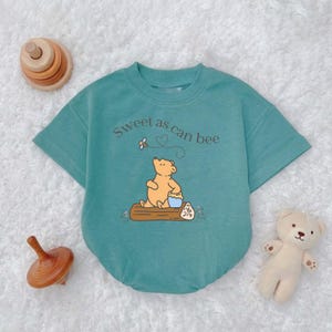 May include: A teal baby romper with short sleeves and the phrase "Sweet as can bee" printed on the front. The romper features a cartoon bear illustration sitting on a log with a honey pot. Wooden toys and a stuffed bear are also in the image.