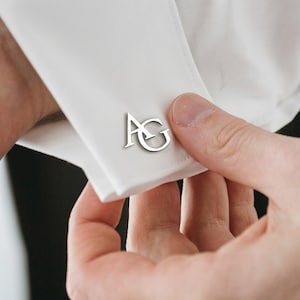 Op de afbeelding: Zilveren manchetknopen met de initialen "AG" worden getoond op een wit overhemd. De manchetknopen, met een gepolijste, reflecterende afwerking, staan centraal. De afbeelding benadrukt de details van het overhemd en de manchetknopen.