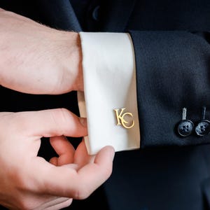 Peut inclure: Boutons de manchette dorés avec les initiales "KC" sur un poignet de chemise blanche. Le poignet est mis en valeur sur une veste de costume bleu marine foncé. L'image est un gros plan du poignet et de la main.
