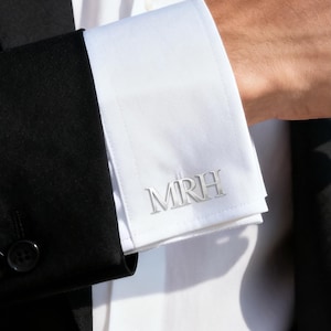 Puede incluir: Gemelos plateados con las iniciales MRH en una camisa blanca. El gemelo es visible contra una chaqueta de traje negra. La imagen es un primer plano del gemelo.