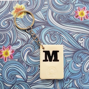 May include: A cream-colored keychain with a black letter "M" on a white background. The keychain is attached to a silver chain and ring.