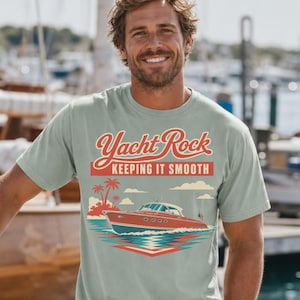 May include: A light green t-shirt with the text "Yacht Rock KEEPING IT SMOOTH" and a graphic of a red boat on the water. The shirt is worn with khaki shorts. The background shows a marina and a person.