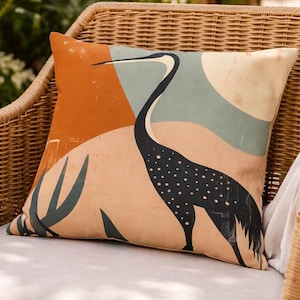 Könnte beinhalten: Quadratisches Zierkissen mit Reiher-Design. Das Kissen zeigt einen schwarzen Reiher mit weißen Tupfen vor einem Hintergrund aus orangefarbenen, beigen und türkisfarbenen Formen. Das Kissen liegt auf einem Korbstuhl.